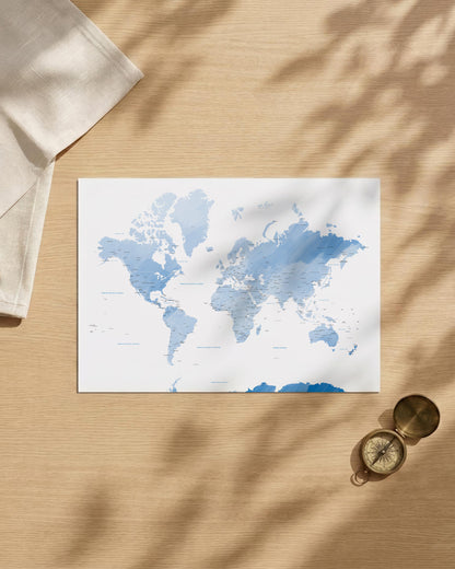 A watercolor world map print in shades of blue on a white background, styled in a flat-lay on a light wood surface with leaf shadows, a folded cream cloth, and a brass compass.