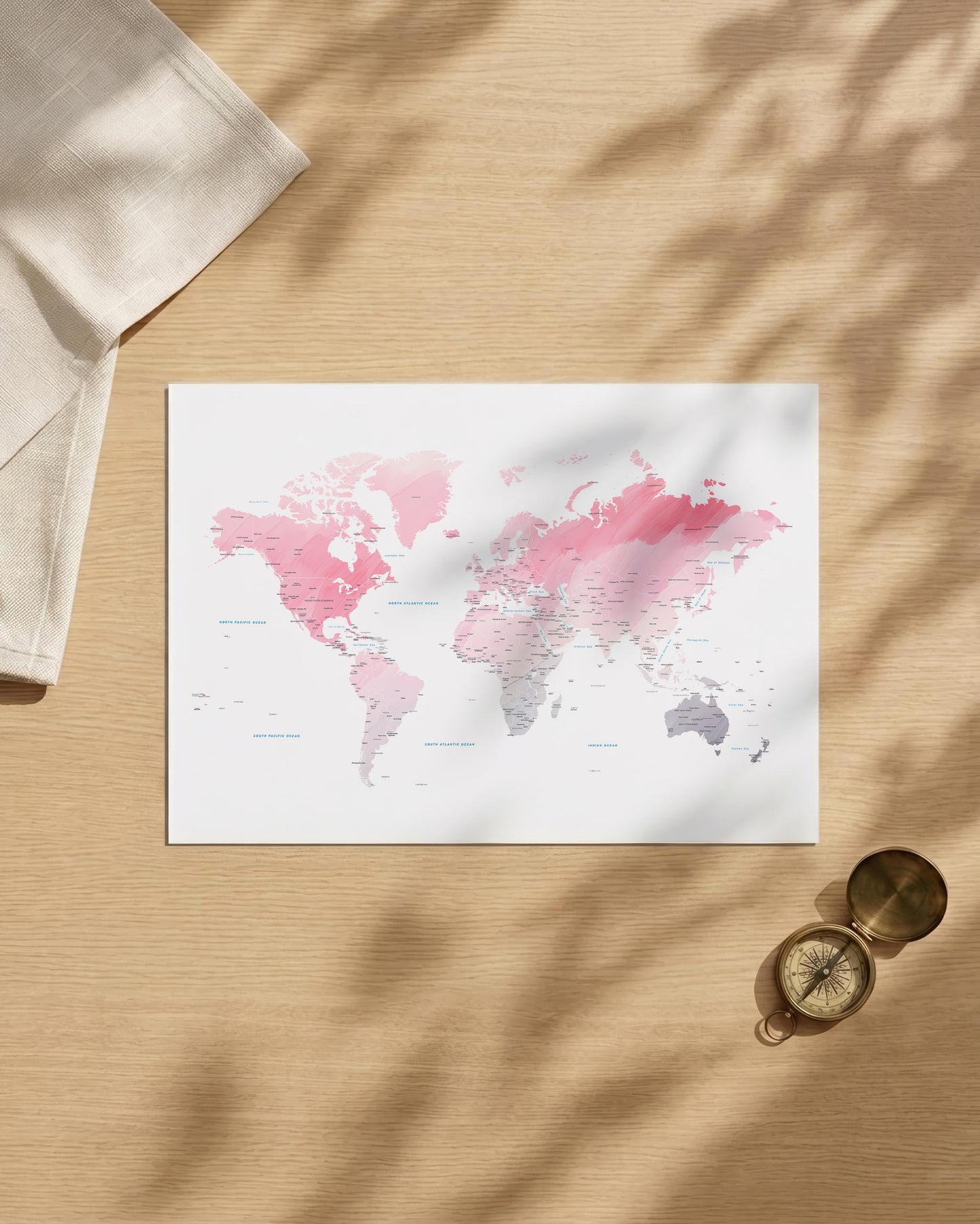 A world map art print with pink and gray watercolor continents on a white background, styled in a flat lay on a light wood surface with leaf shadows, a folded cream cloth, and a brass compass.