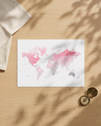 A world map art print with pink and gray watercolor continents on a white background, styled in a flat lay on a light wood surface with leaf shadows, a folded cream cloth, and a brass compass.