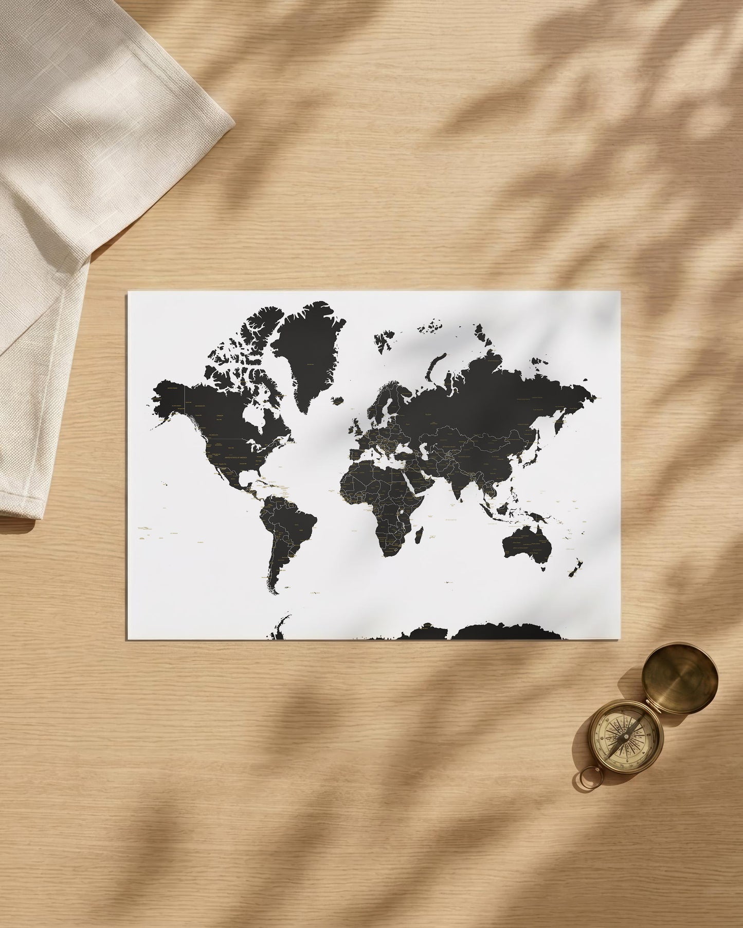 A rectangular world map art print with black landmasses on a white background, styled on a light wood surface with leaf shadows, a folded cream cloth, and a small brass compass.
