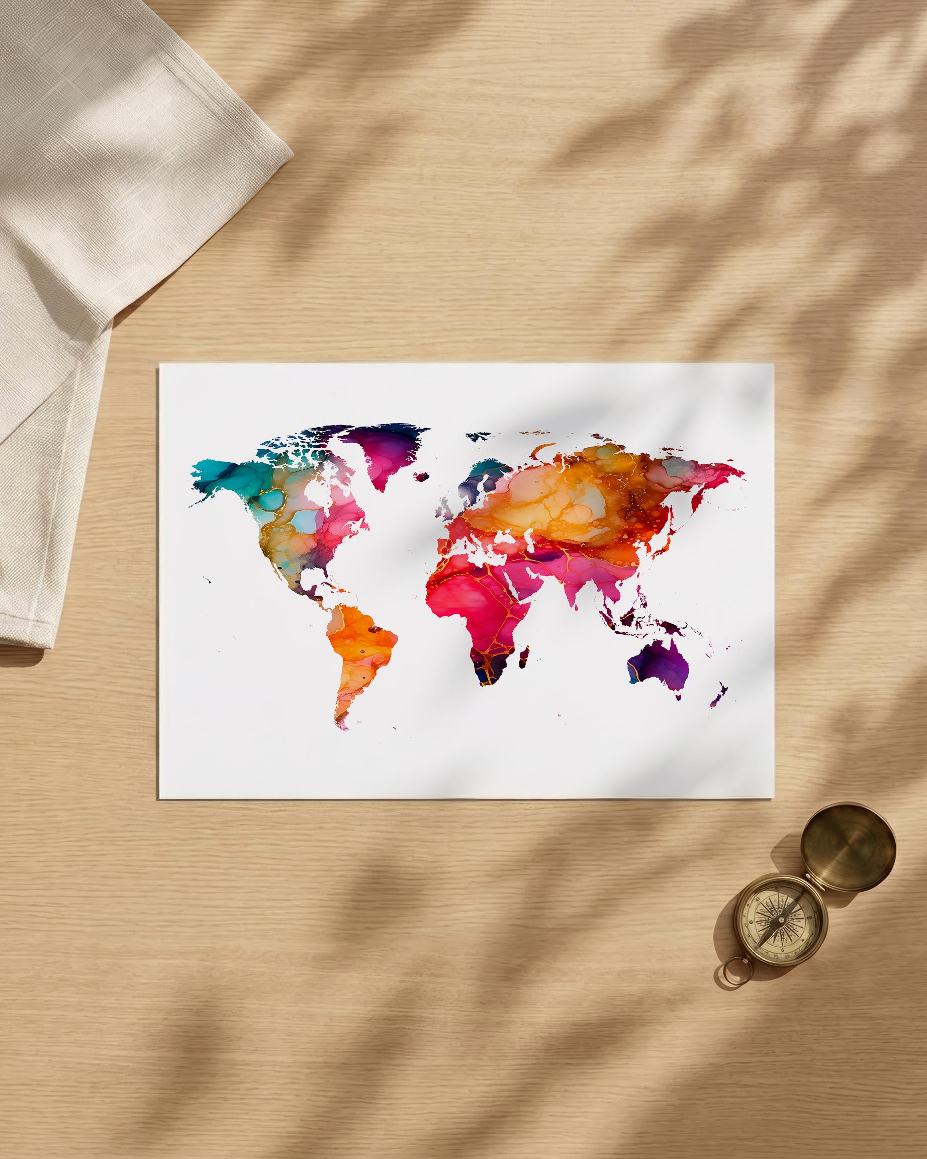 A rectangular art print featuring a colorful watercolor world map on a white background, styled on a light wood surface with soft leaf shadows, a folded cream cloth, and a small brass compass.
