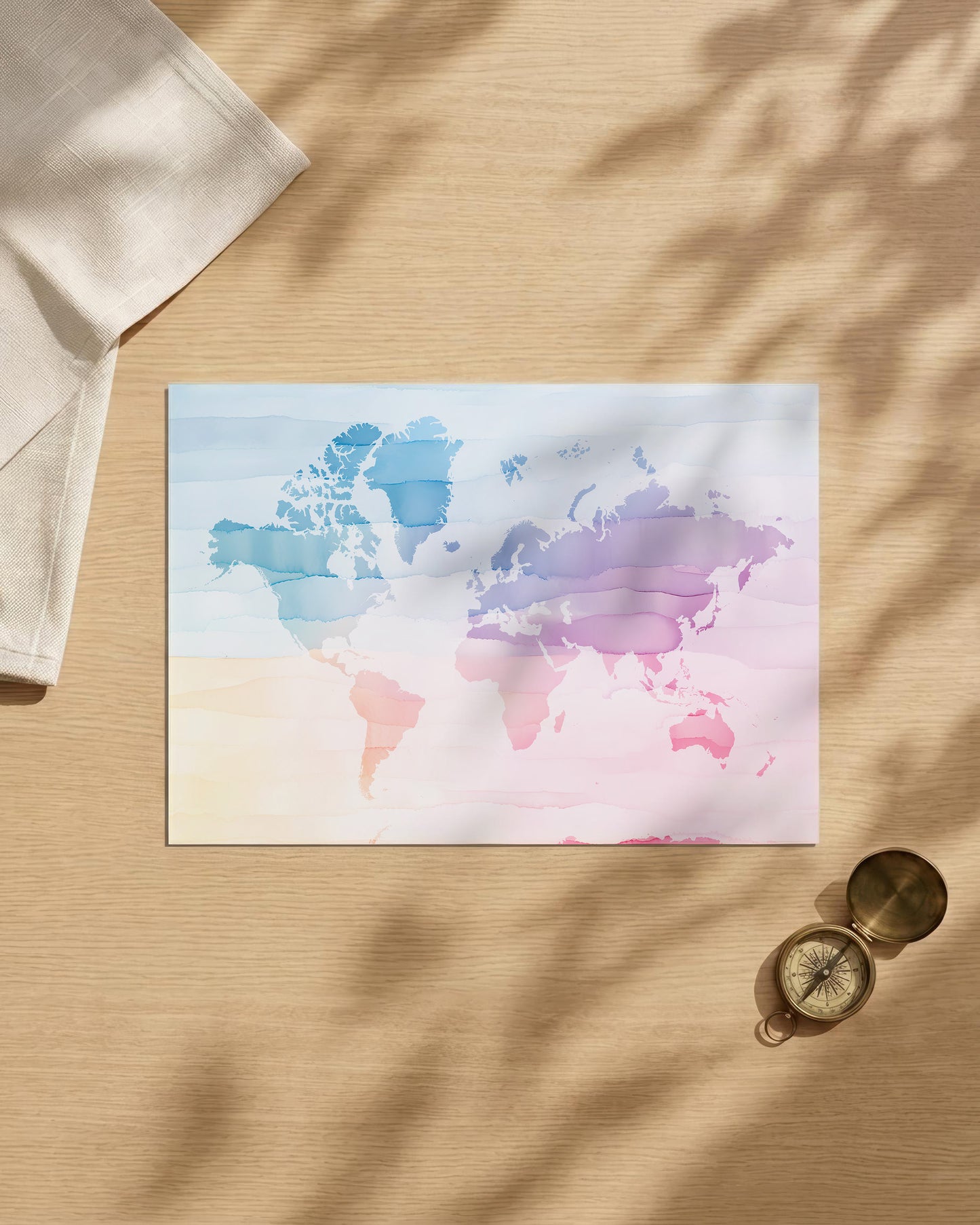 A watercolor world map art print with pastel blue, purple, and pink continents, styled on a light wood surface with soft leaf shadows, a folded cream cloth, and a small brass compass.