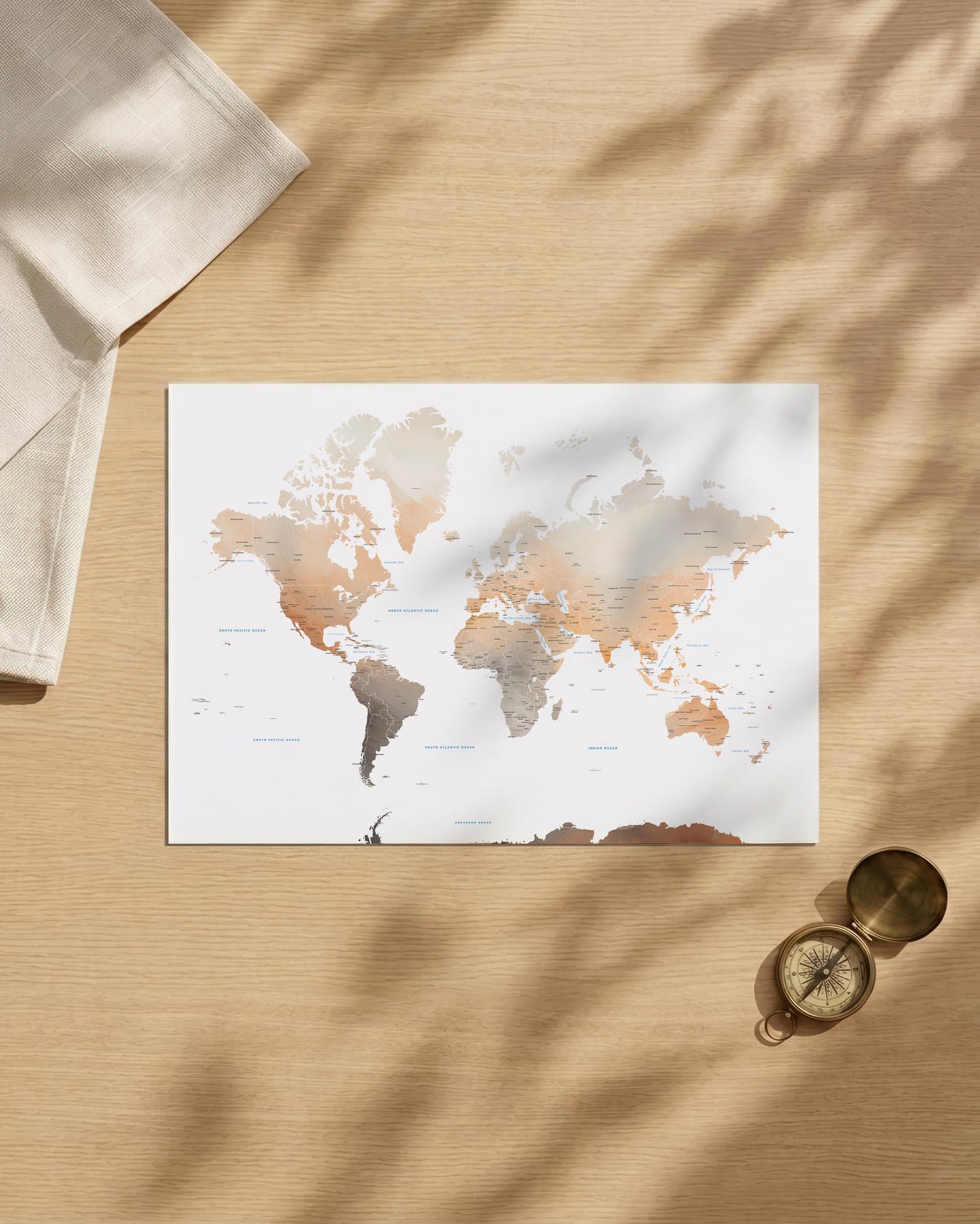 A world map art print with a watercolor finish in neutral beige, brown, and gray tones, styled in a flat-lay on a light wood surface with leaf shadows, a folded cream cloth, and a brass compass.