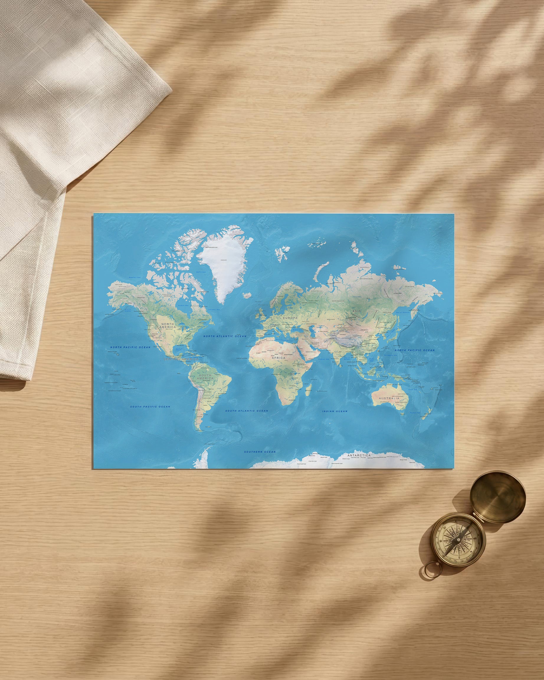 A rectangular world map print with a blue ocean background and pale landmasses, styled on a light wood surface with soft leaf shadows, a folded cream cloth at upper left, and a brass compass at lower right.