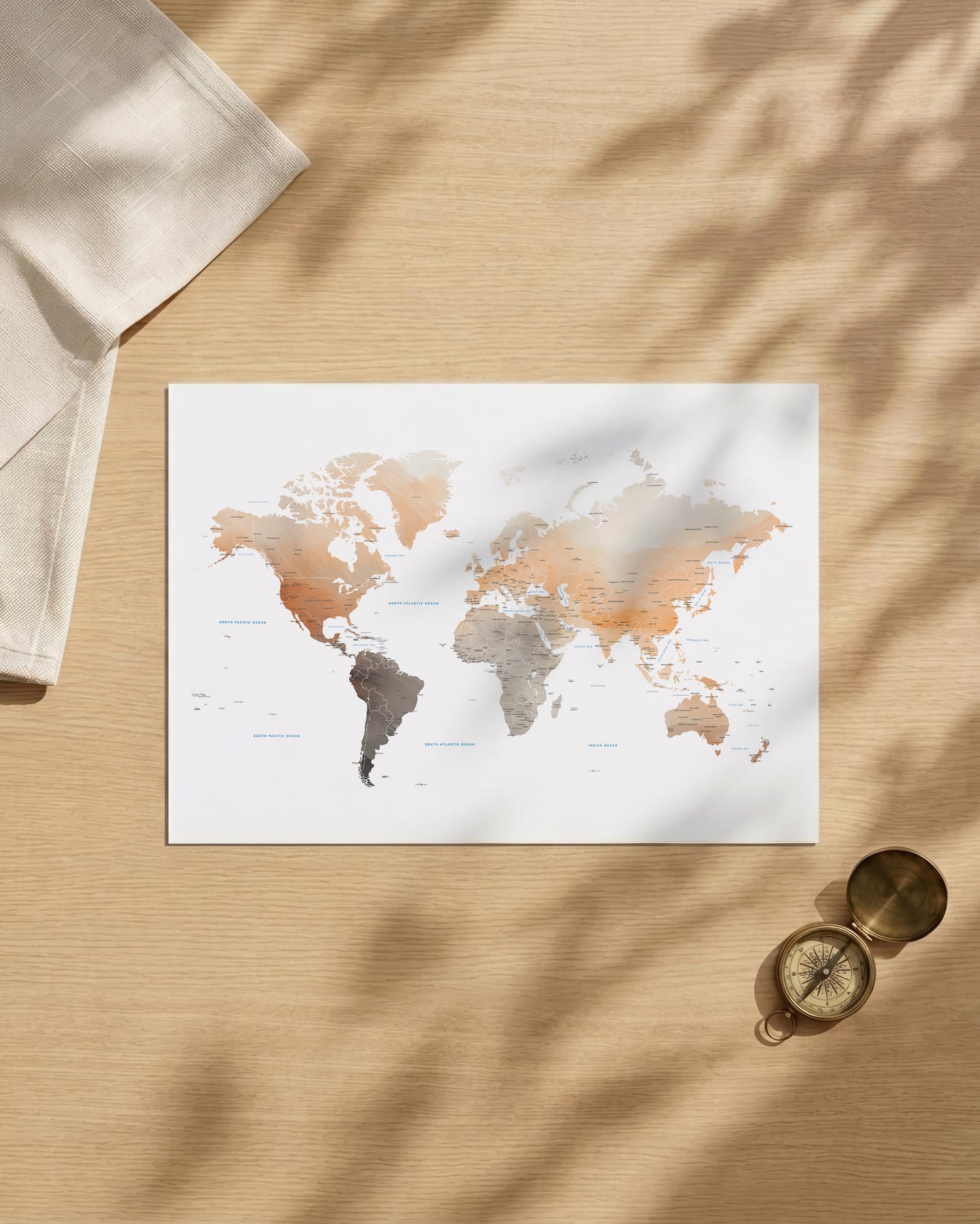 A rectangular world map art print with a watercolor-style finish in muted beige, brown, gray, and white tones, displayed on a light wood surface with soft leaf shadows. A folded cream cloth and a small brass compass are placed nearby as styled props.