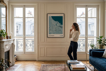A woman stands in a bright, elegant living room looking at a framed poster hanging on a cream paneled wall. The poster features a teal and white map design labeled 'CHAMBERY'.