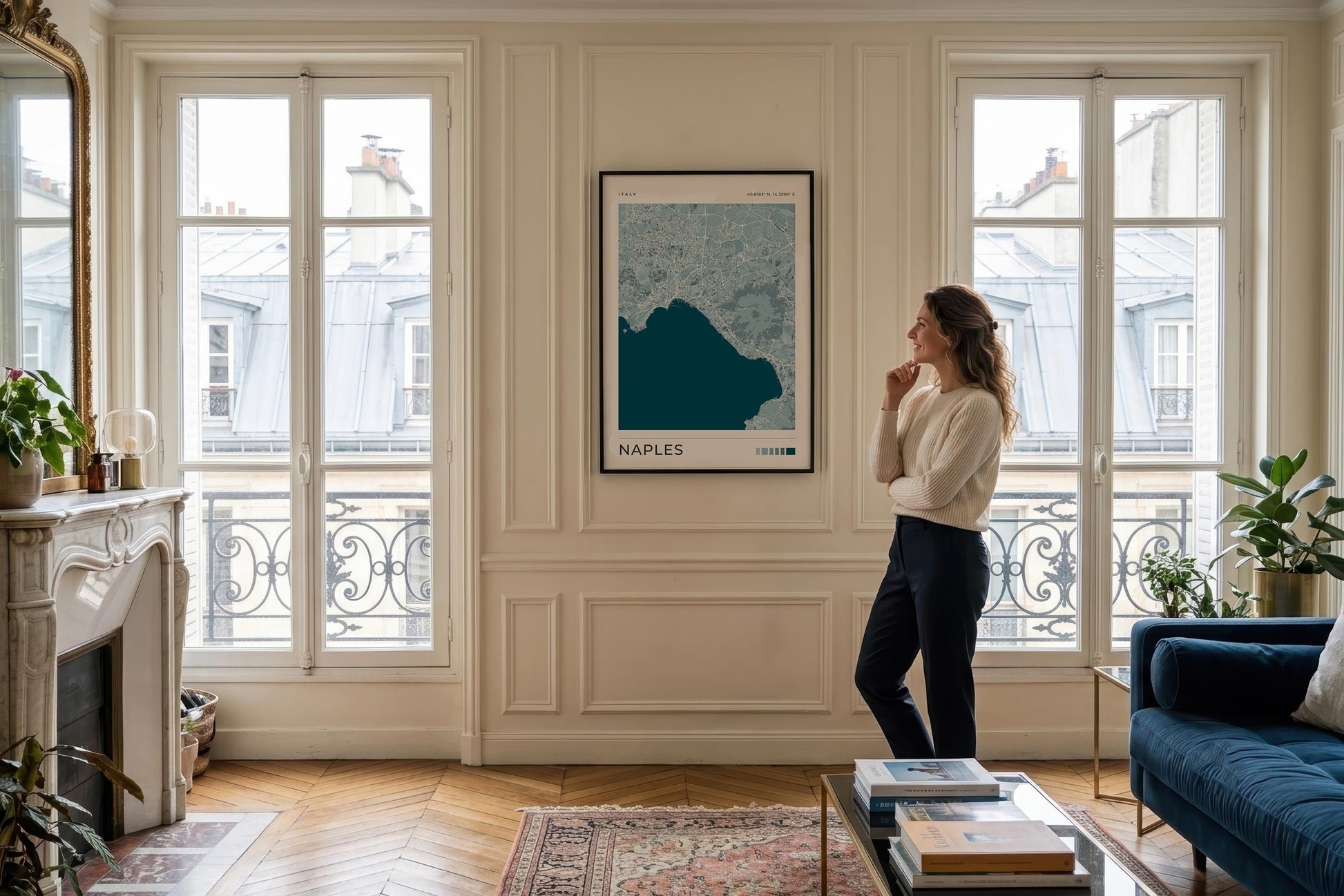 A woman stands in a bright, elegant living room looking at a framed Naples map art print hanging on a cream paneled wall. The room features classic French windows, a fireplace, a blue velvet sofa, and a wooden parquet floor.