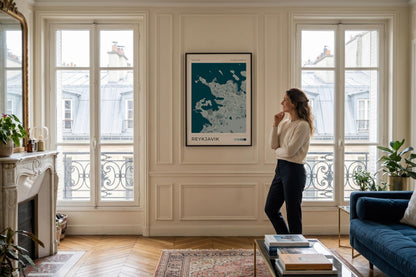 A woman stands in a bright, elegant room looking at a framed map print of Paris on the wall. The room features classic cream paneled walls, large multi-paned windows looking out onto city buildings, a fireplace on the left, and a blue sofa on the right.