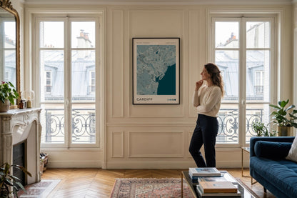 A woman stands in a bright, elegant living room looking at a framed map print titled 'CARDIFF' hanging on a cream paneled wall between large windows.