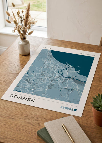A stylized flat-lay of a Gdańsk city map poster on a wooden table, surrounded by a small vase of dried grass, a succulent, and a notebook with a pen.