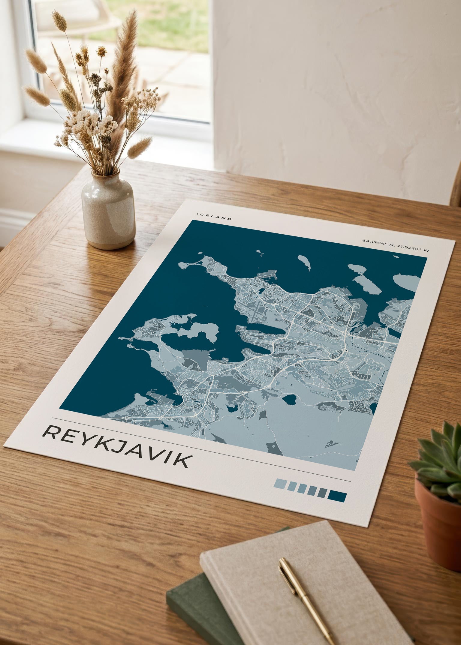 A minimalist Reykjavík map art print displayed on a wooden desk next to a small vase of dried grass, a succulent, and a notebook with a pen.