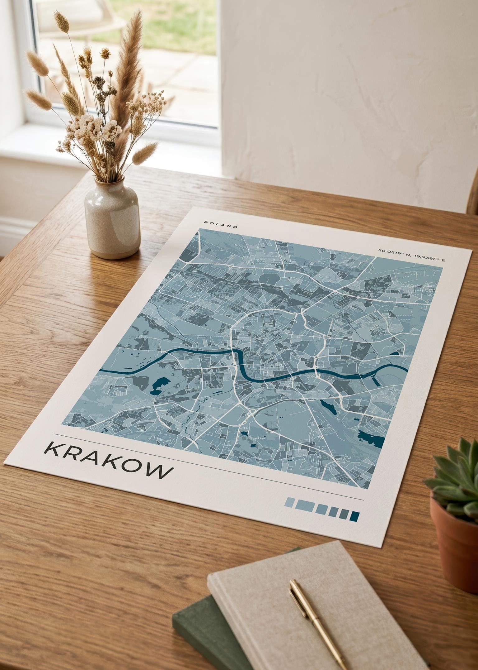 A blue-toned Krakow city map print displayed on a wooden table, styled with a small vase of dried grasses, a notebook, and a succulent.