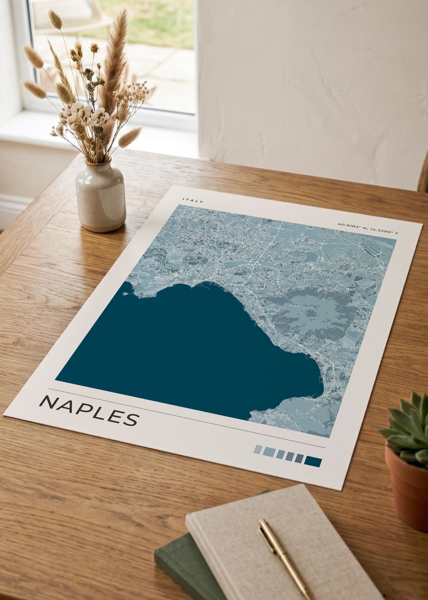 A printed Naples cityscape art poster with a dark teal sea and light blue/gray land lies on a wooden table, styled with a small vase of dried grass, a succulent, and notebooks nearby.