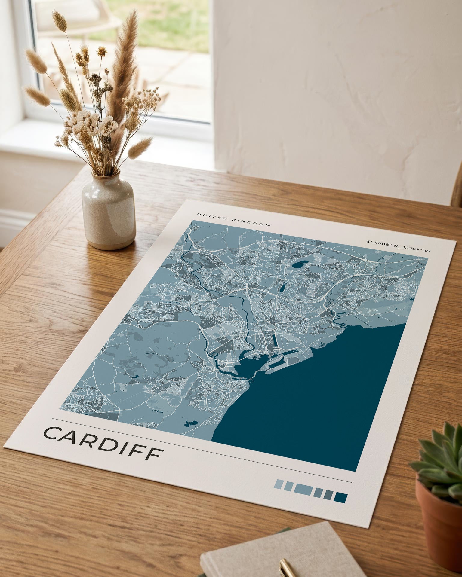 A city map print of Cardiff, United Kingdom, laid on a wooden table in a bright interior. The print shows a blue and white cartographic layout with the title 'CARDIFF' and 'UNITED KINGDOM' at the top.