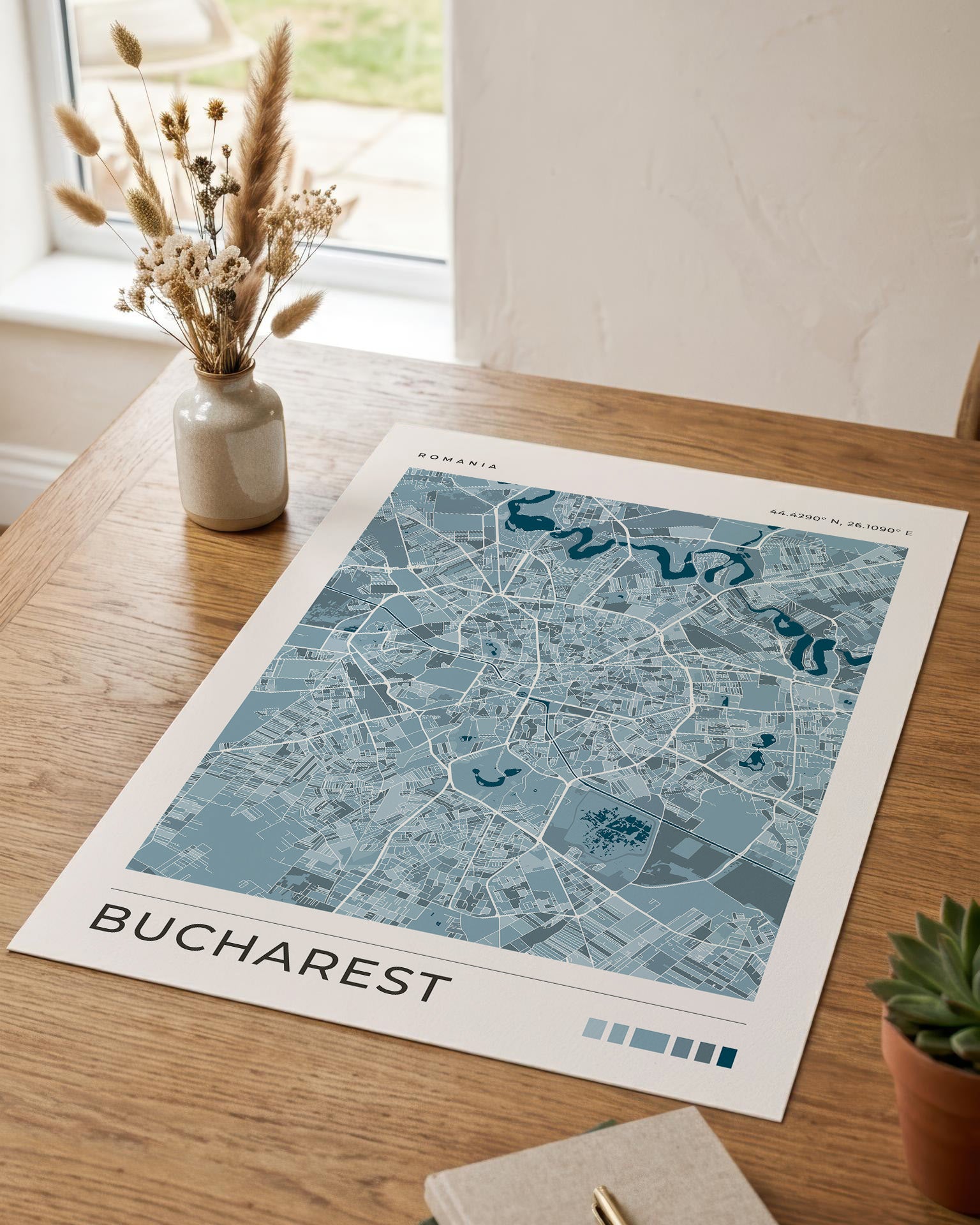 A blue-toned street map print of Bucharest, Romania, displayed on a wooden table in a styled interior setting with a vase of dried grasses, a notebook, and a small succulent.