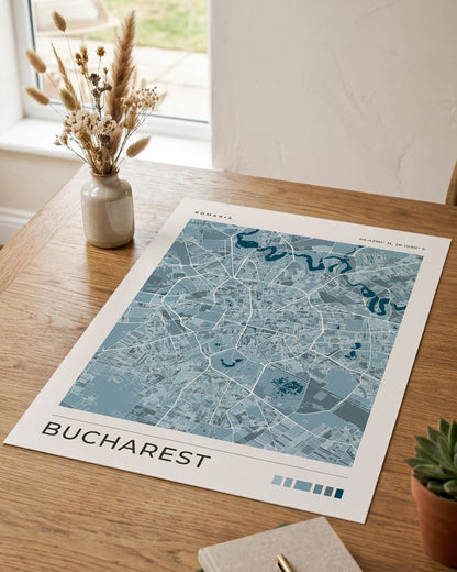 A blue-toned street map print of Bucharest, Romania, displayed on a wooden table in a styled interior setting with a vase of dried grasses, a notebook, and a small succulent.