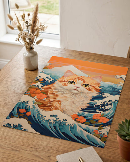 A rectangular art print or poster lying on a wooden table, featuring a stylized orange-and-white cat in a blue ocean scene with waves and flowers, inspired by Japanese wave art. The print is styled in a real indoor setting with a vase of dried grasses, a notebook, and a small plant nearby.