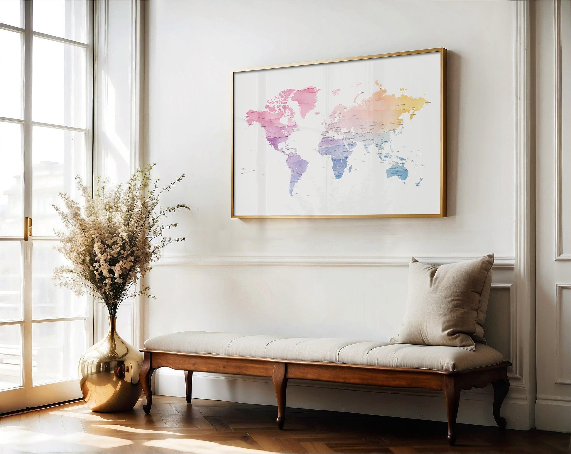 A framed world map art print with a gold frame hangs on a white wall above a wooden bench with a beige cushion in a bright, sunlit room.