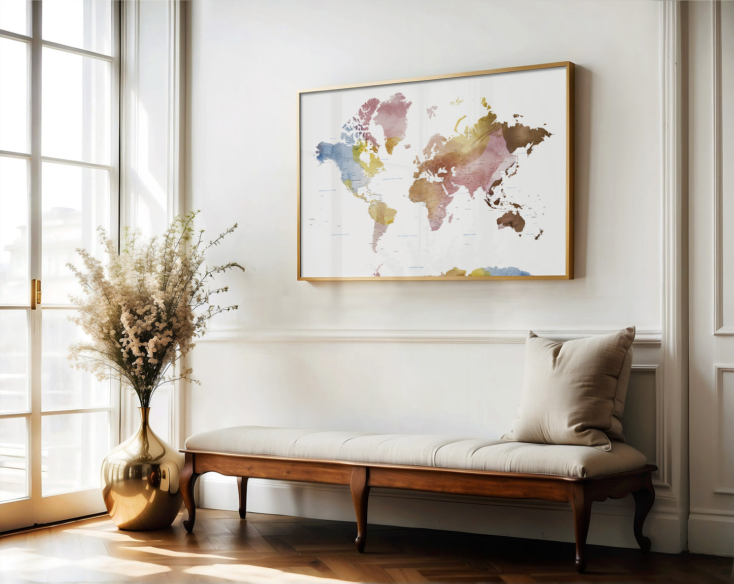 A framed world map wall art print hangs above a wooden bench with a beige cushion in a bright, modern room with a window and a vase of dried flowers.