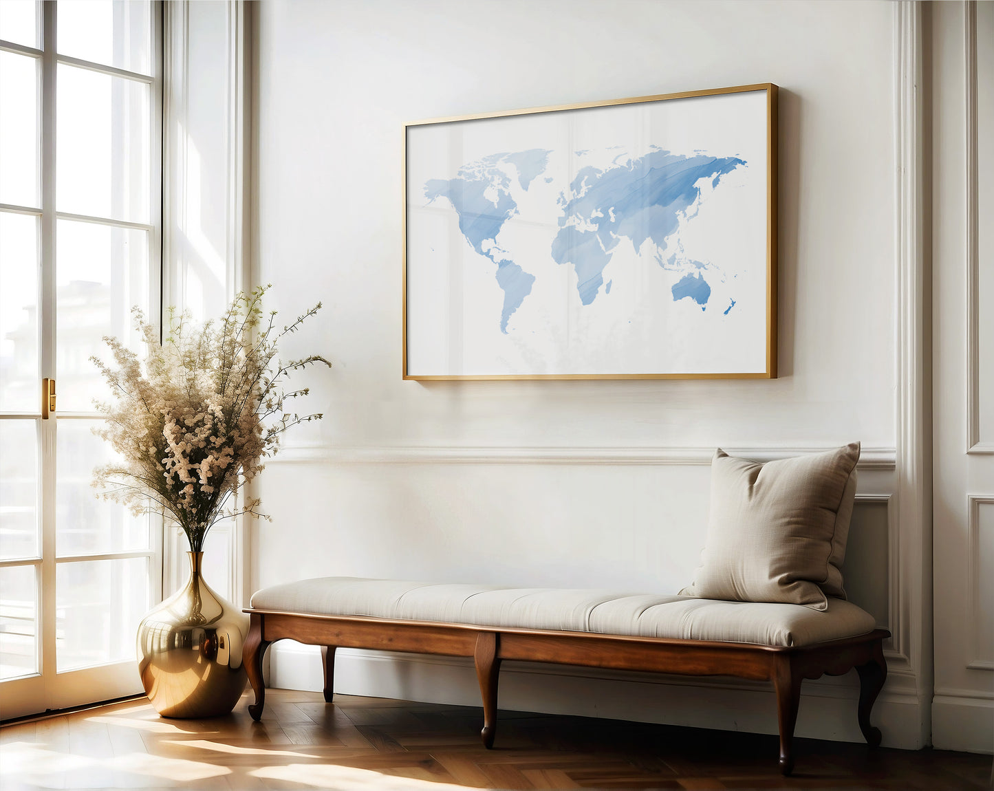 A framed world map art print in light blue watercolor tones with a gold frame, displayed on a white wall above a wooden bench with a beige cushion in a bright, sunlit room.