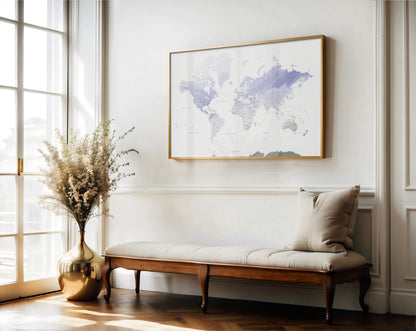 A framed world map art print with a light wood frame hangs on a white wall above a wooden bench with a beige cushion in a bright, sunlit room.