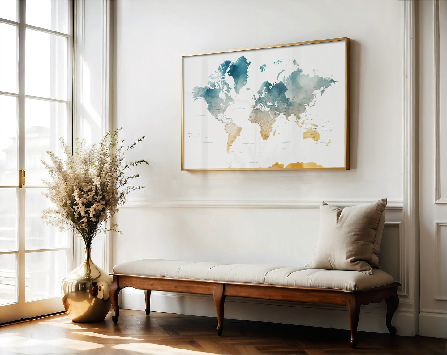 A framed world map art print with blue, green, and gold watercolor details hangs on a white wall above a wooden bench with a beige cushion in a bright, modern room.
