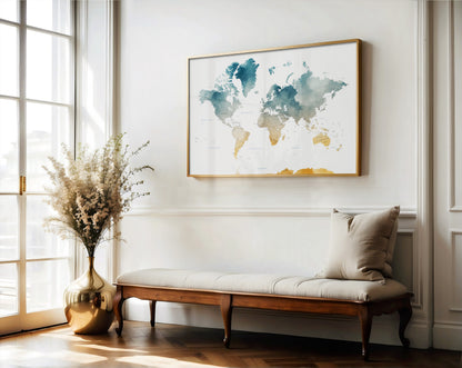 A framed world map art print with blue, green, and gold watercolor details hangs on a white wall above a wooden bench with a beige cushion in a bright, modern room.