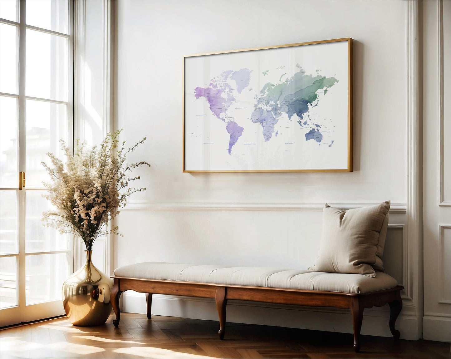 A framed world map art print with watercolor-style continents in purple, blue, and green tones, hanging on a white wall above a wooden bench with a cushion in a bright, sunlit room.