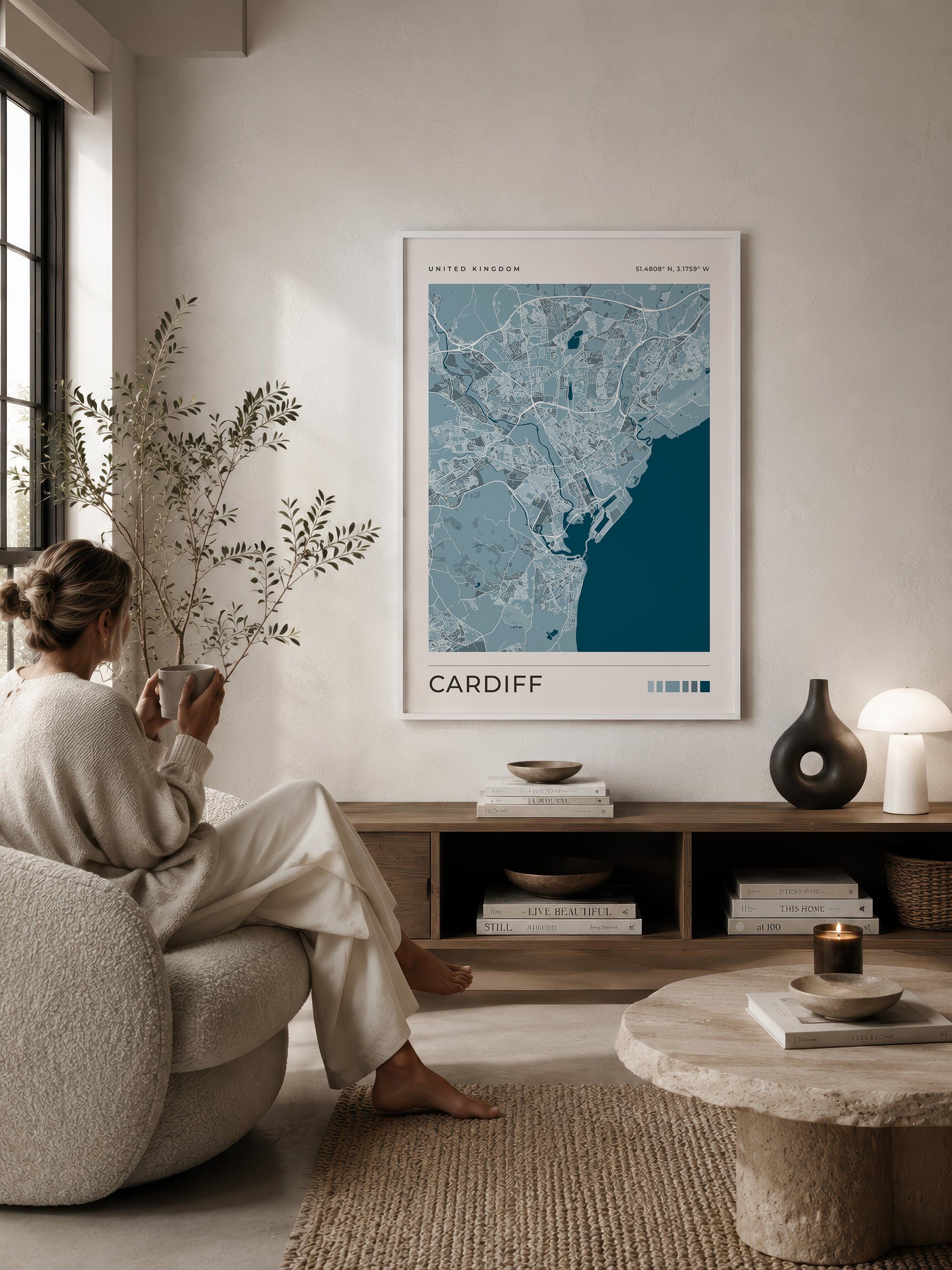 A framed map print of Cardiff, Wales, hangs on a beige wall in a modern, sunlit living room. A woman in a cream lounge set sits in a textured armchair, reading a book, while a stone coffee table and wooden shelf with books and decor sit below.