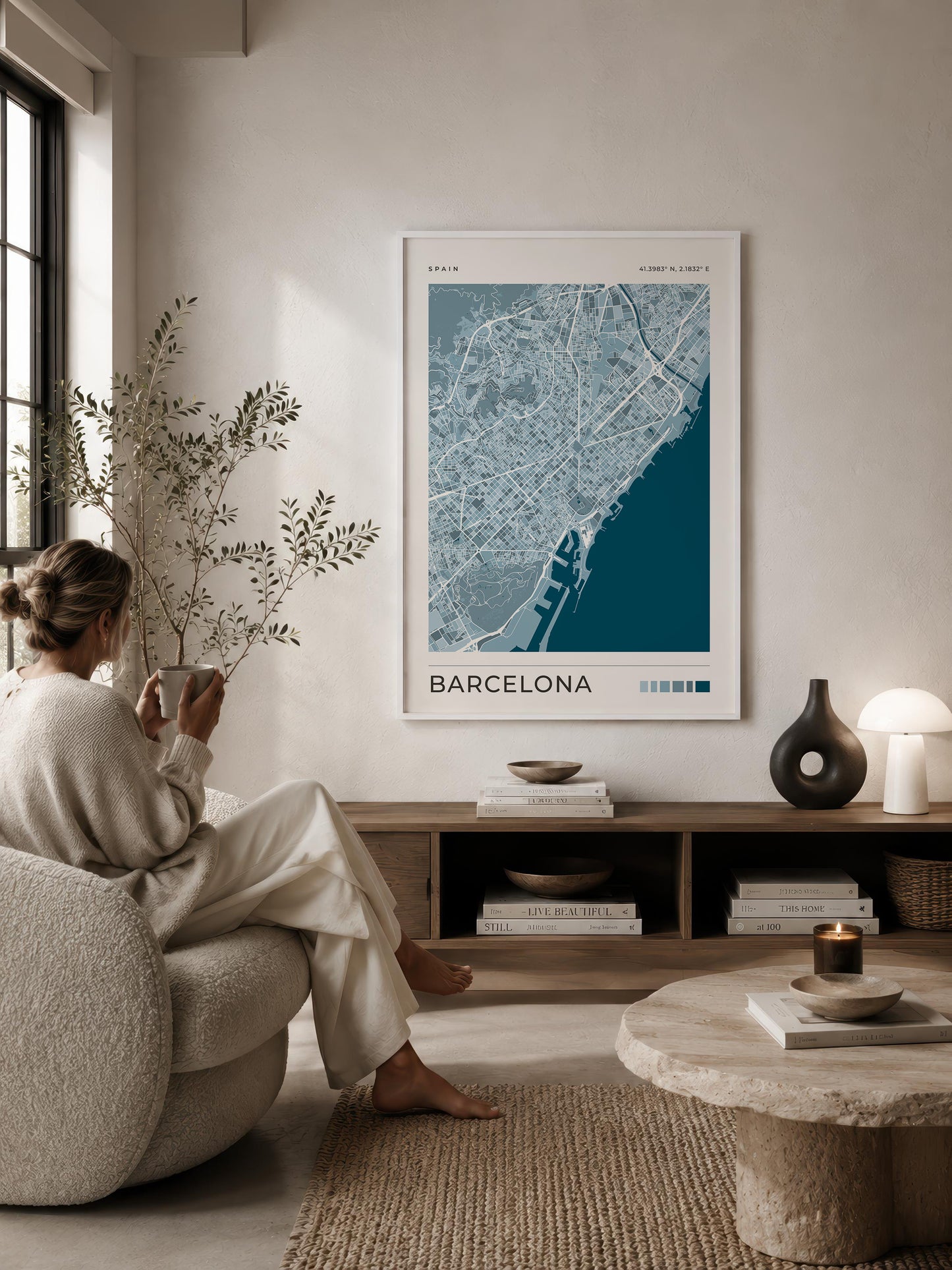 A framed Barcelona city map poster hangs on a beige wall in a styled living room. A woman sits in a textured armchair facing the artwork, with a wooden console, books, a lamp, and a stone coffee table in the room.