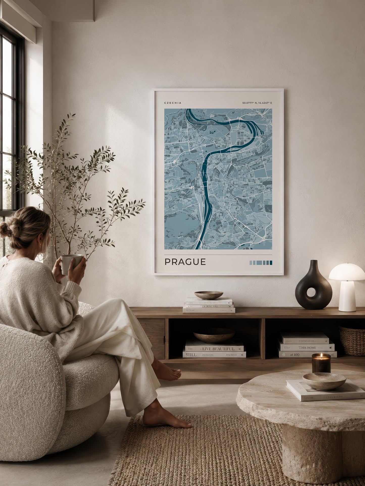A framed blue and white map poster of Prague hangs on a beige wall in a modern, sunlit living room. A woman in a cream lounge set sits on a textured armchair to the left, and a wooden console table with books and decor is below the poster.