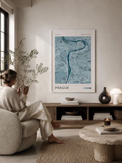 A framed blue and white map poster of Prague hangs on a beige wall in a modern, sunlit living room. A woman in a cream lounge set sits on a textured armchair to the left, and a wooden console table with books and decor is below the poster.