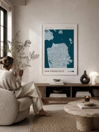 A framed San Francisco city map poster hangs on a beige wall in a styled living room. A woman in a cream robe sits on a textured armchair facing the console, with a stone coffee table, books, and decor in the foreground.