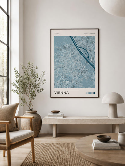 A framed Vienna city map poster hangs on a light gray wall in a modern, sunlit living room with a chair, bench, lamp, and round table.