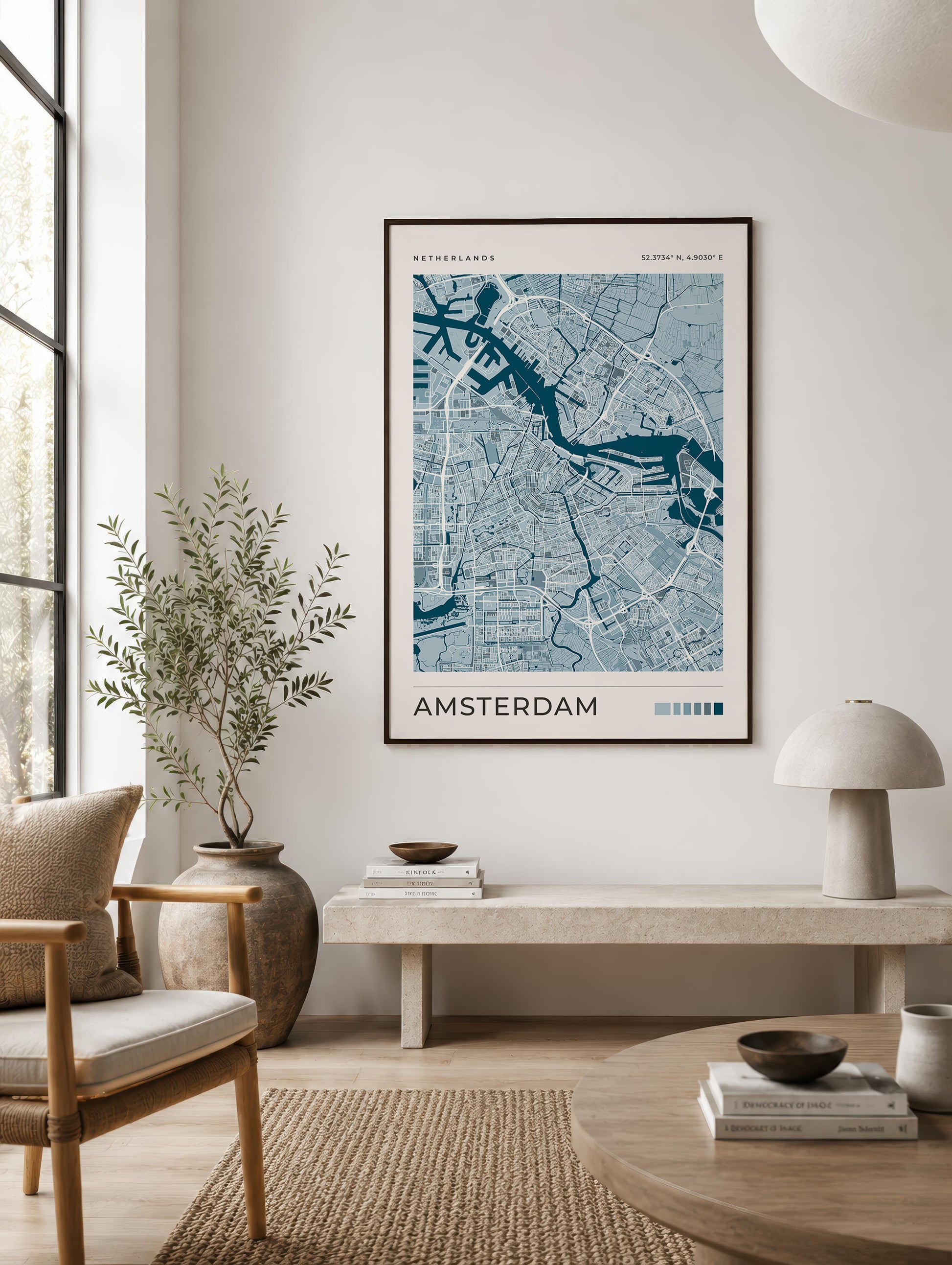 A framed Amsterdam city map poster hangs on a light beige wall in a modern, sunlit living room with a chair, bench, lamp, and rug.