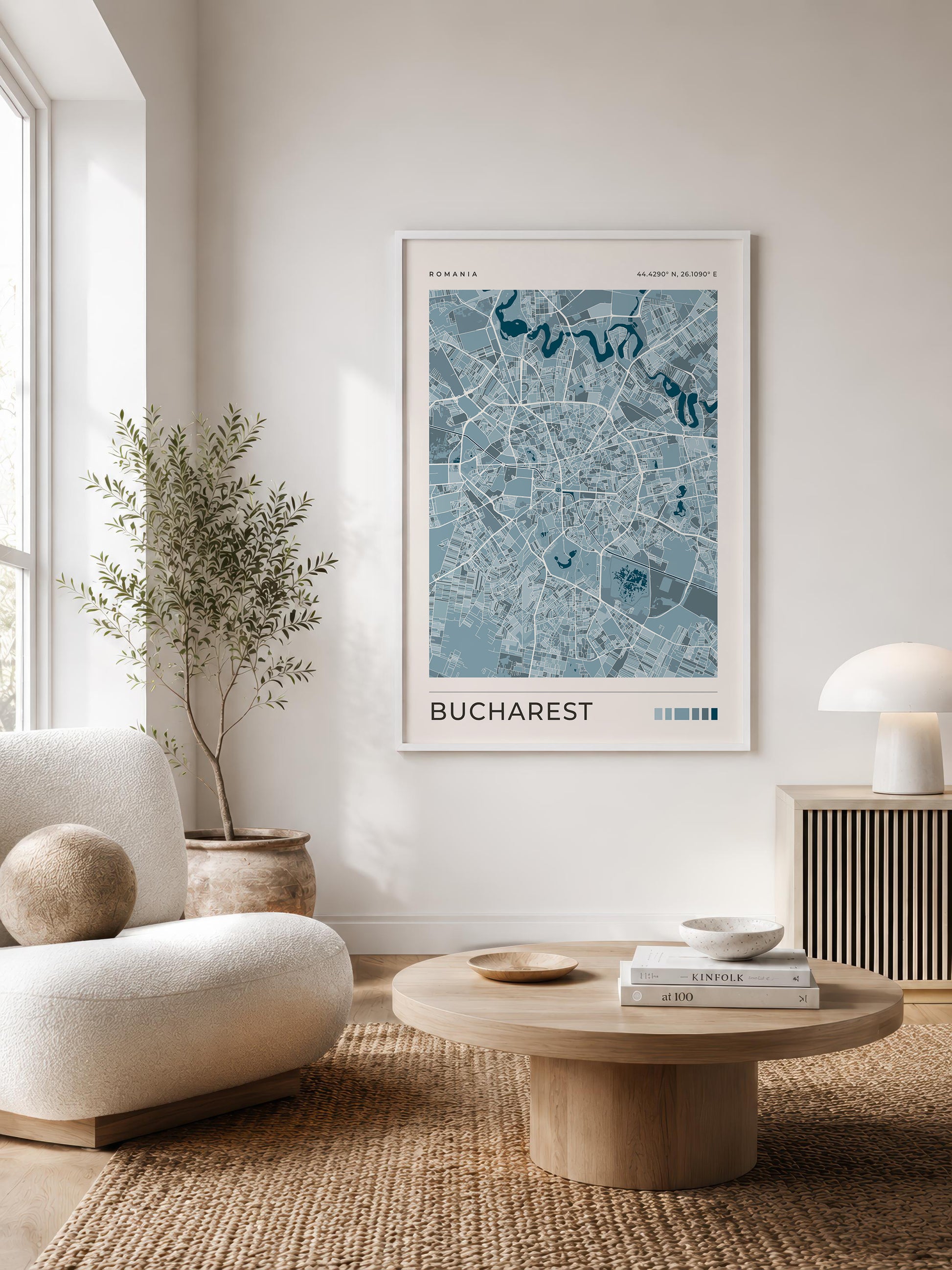 A framed art print of a blue and white map of Bucharest, Romania, styled on a white wall in a modern, sunlit living room with a beige armchair, round wooden coffee table, and potted plant.