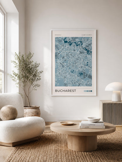 A framed art print of a blue and white map of Bucharest, Romania, styled on a white wall in a modern, sunlit living room with a beige armchair, round wooden coffee table, and potted plant.