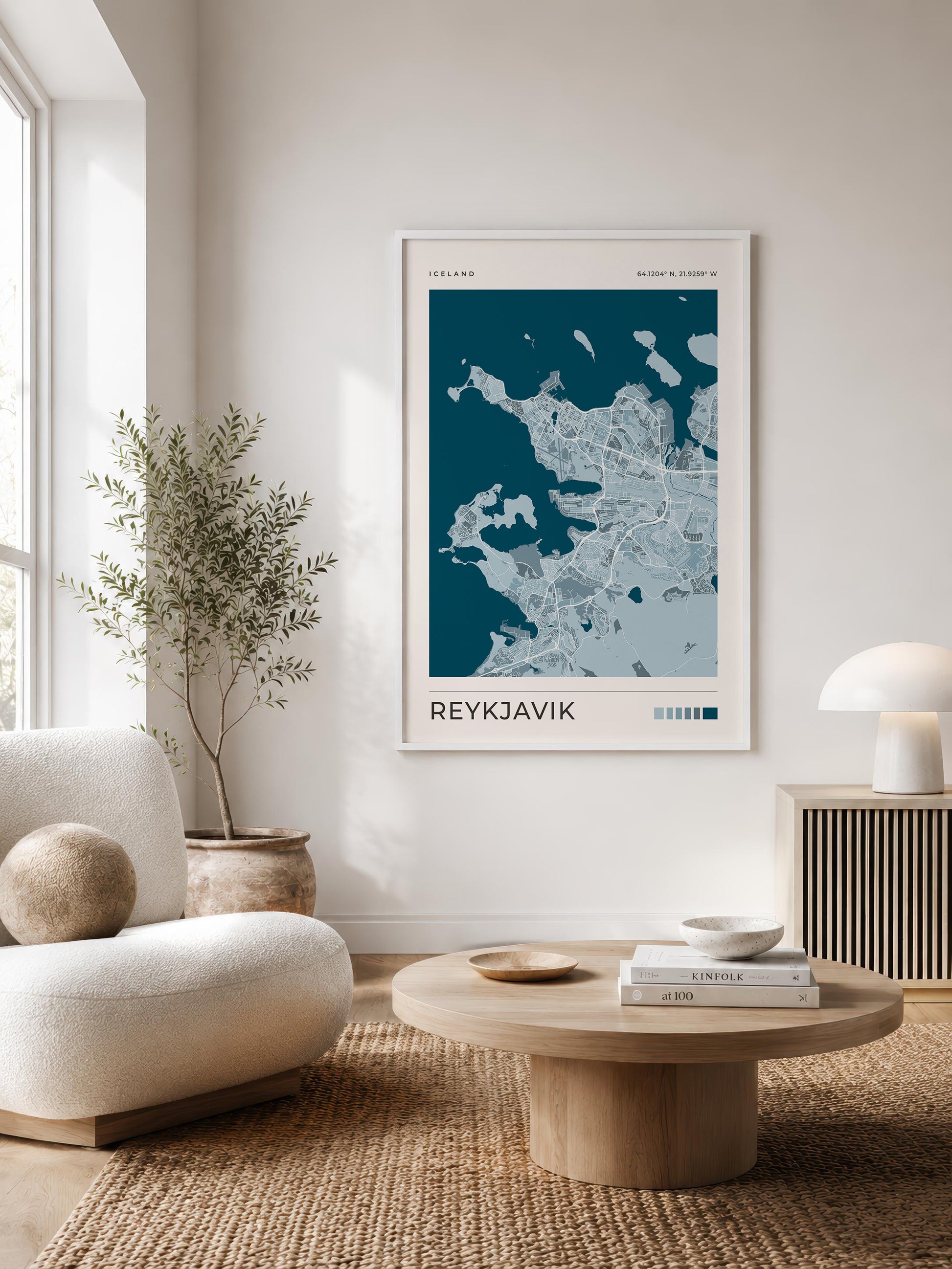 A framed minimalist map print of Reykjavík hangs on a white wall in a modern, sunlit living room with a cream sofa, round wooden coffee table, and a potted plant.