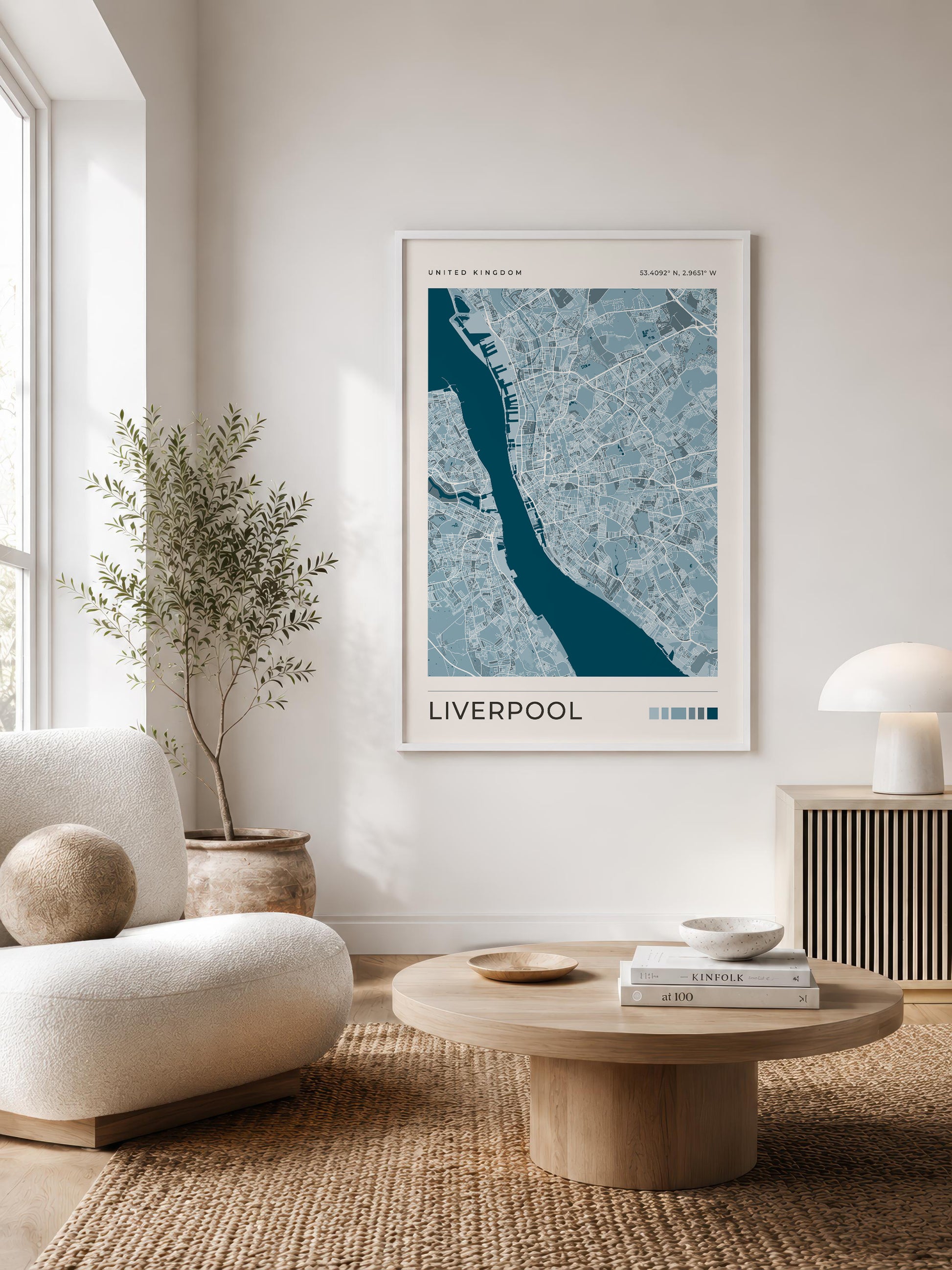A framed art print of a blue and white map of Liverpool, England, styled on a neutral wall in a modern living room setting with a wooden coffee table, a beige armchair, and a potted plant.