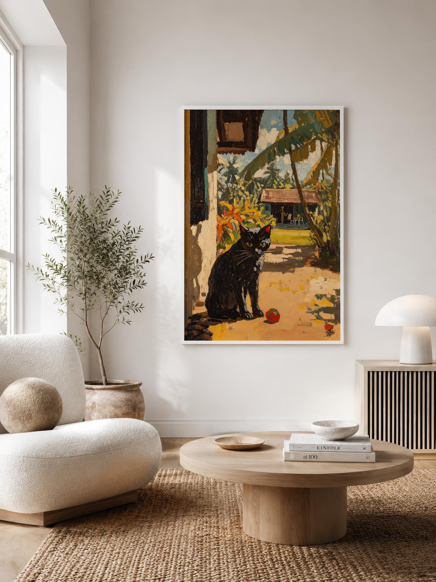 A framed art print depicting a black cat in a tropical outdoor setting, displayed on a white wall in a modern, sunlit living room with a beige armchair, round coffee table, and potted plant.