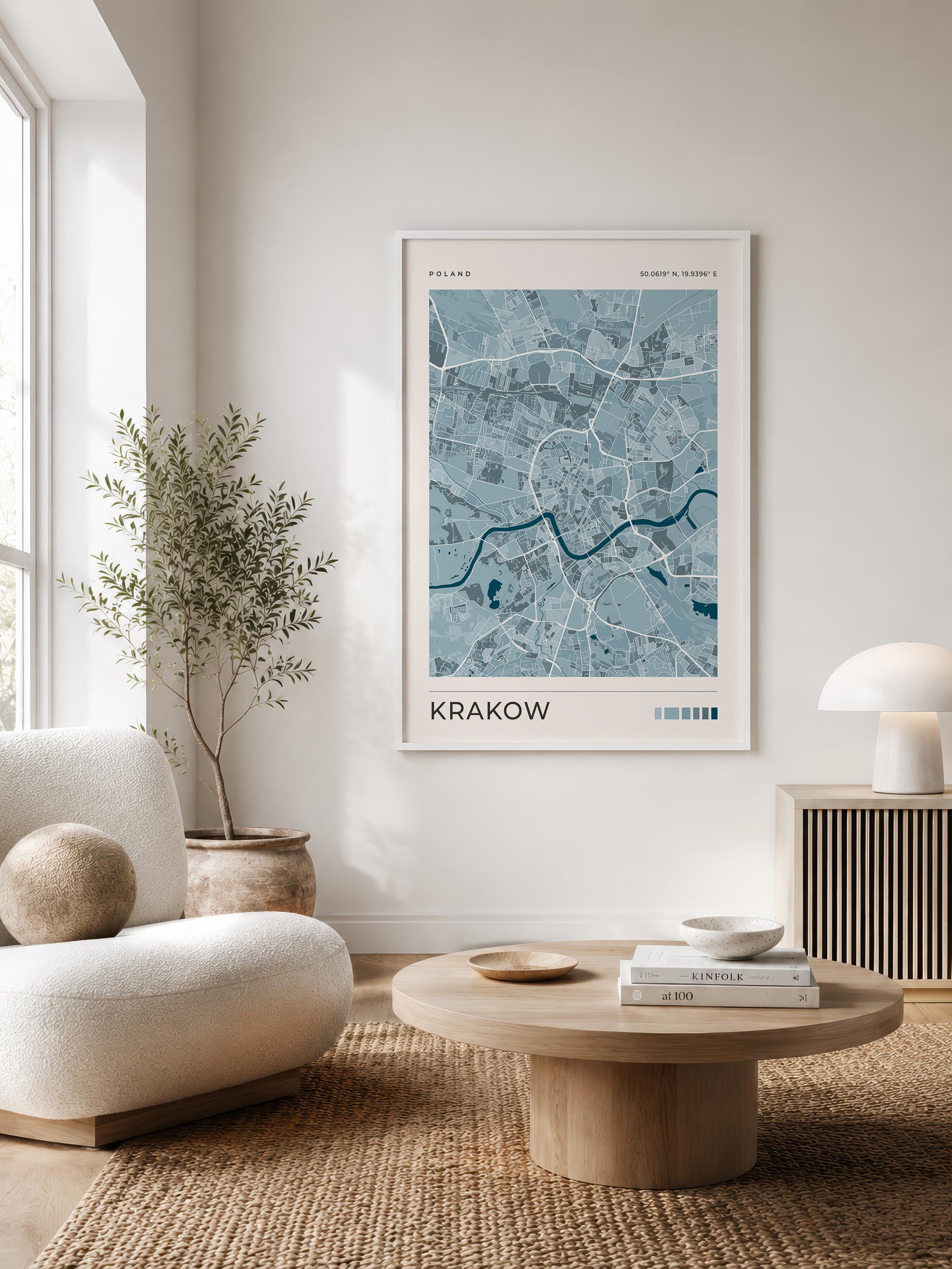 A framed city map print of Krakow, Poland, displayed on a light wall in a modern, sunlit living room with a beige armchair, round wooden coffee table, and a potted plant.
