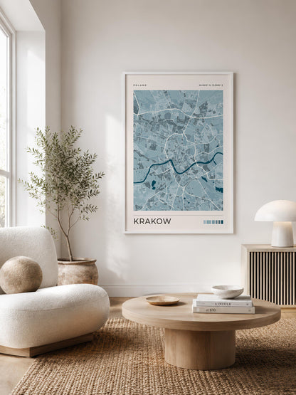A framed city map print of Krakow, Poland, displayed on a light wall in a modern, sunlit living room with a beige armchair, round wooden coffee table, and a potted plant.