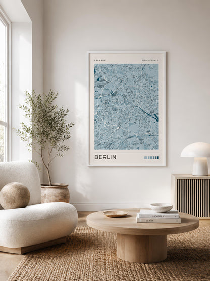 A framed art print of a blue and white Berlin street map hangs on a light beige wall in a modern, sunlit living room with a round wooden coffee table, a cream armchair, and a potted plant.