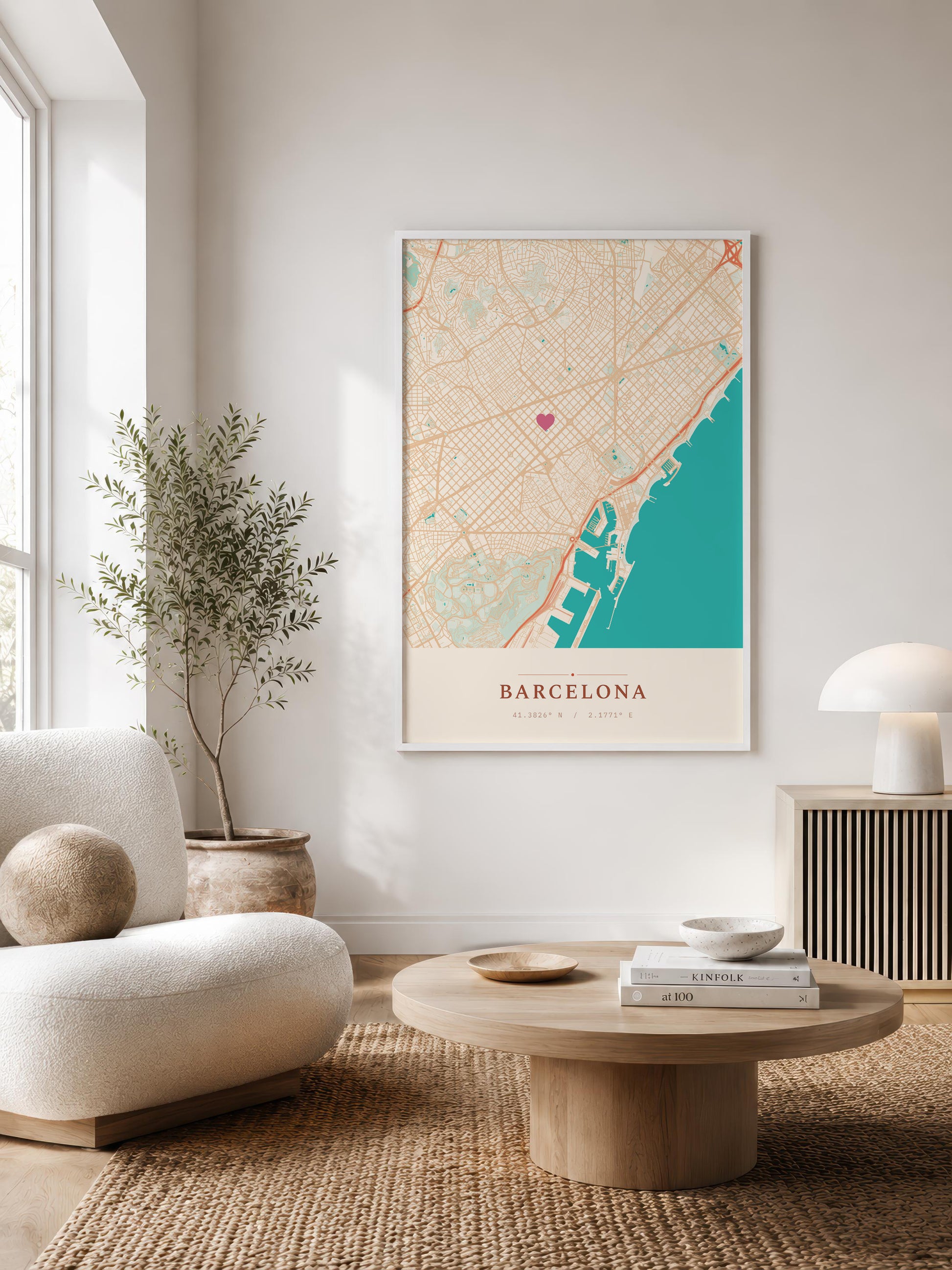 A framed vertical map print of Barcelona, Spain, styled on a neutral wall in a modern living room setting with a beige armchair, round wooden coffee table, and a potted plant.