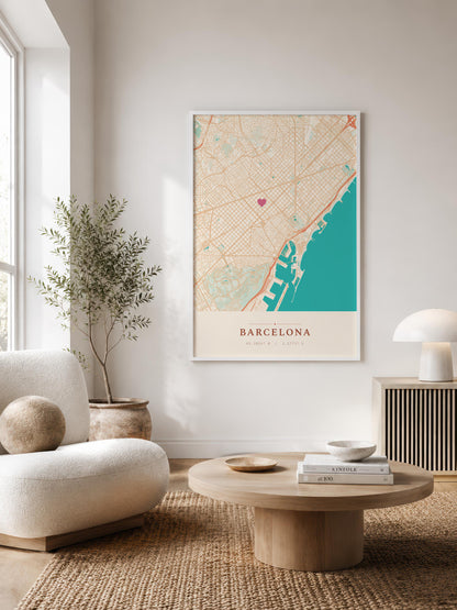 A framed vertical map print of Barcelona, Spain, styled on a neutral wall in a modern living room setting with a beige armchair, round wooden coffee table, and a potted plant.