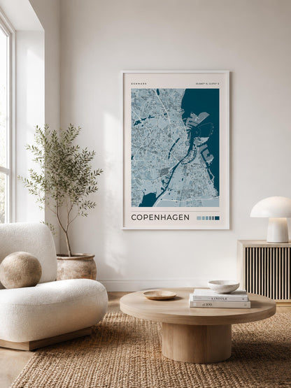A framed Copenhagen city map print in blue and white hangs on a light beige wall in a modern, sunlit living room with a cream sofa, round wooden coffee table, and a potted plant.