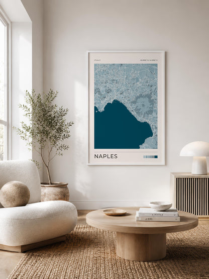 A framed Naples city map art print hangs on a white wall in a modern, sunlit living room with a cream sofa, round wooden coffee table, and a potted plant.