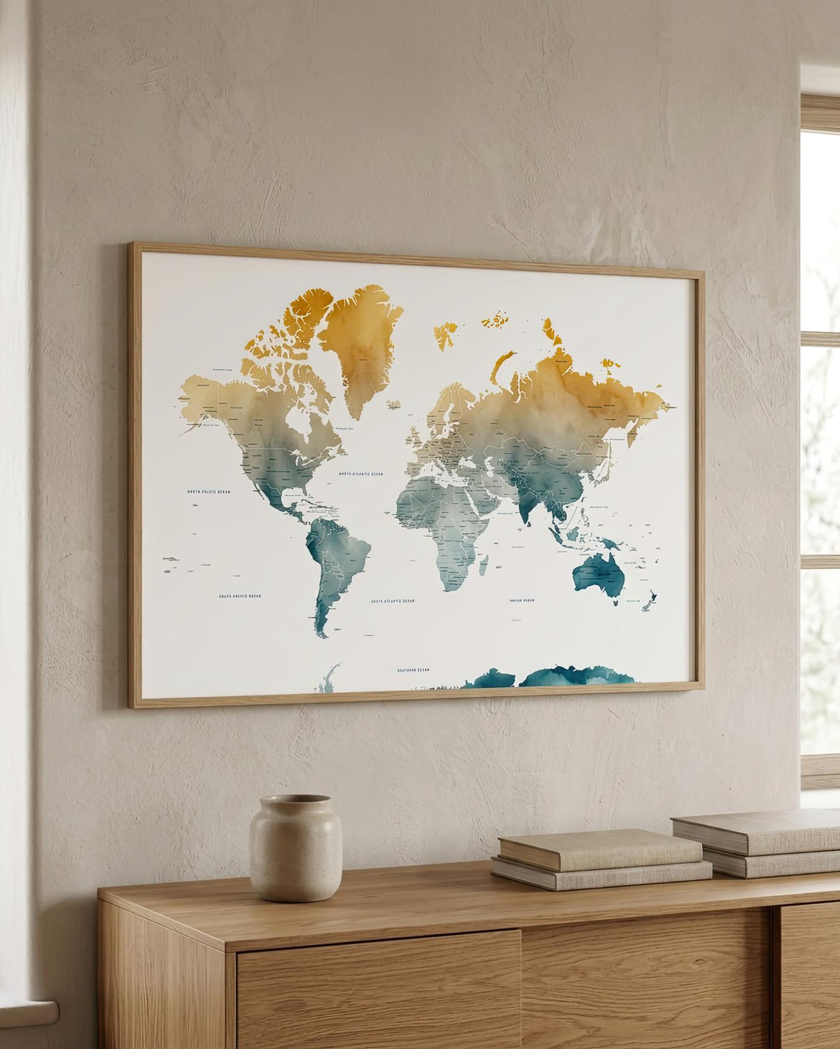 A framed world map art print with watercolor-style continents in gold, teal, and blue on an off-white background, displayed on a beige wall above a light wood console table with books and a small vase.