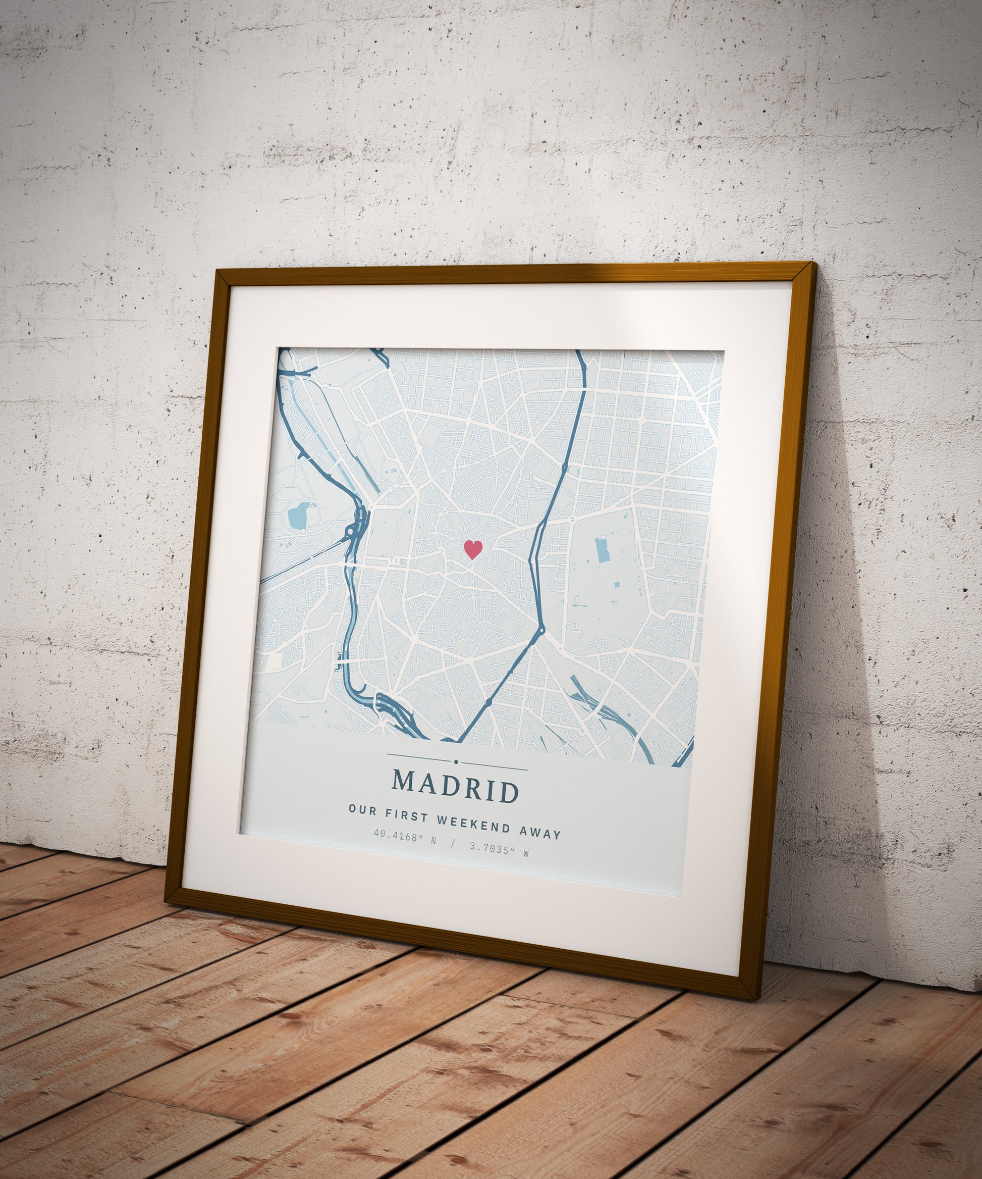A framed personalized Madrid city map print with a gold frame and white mat, leaning against a white brick wall on a wooden floor.