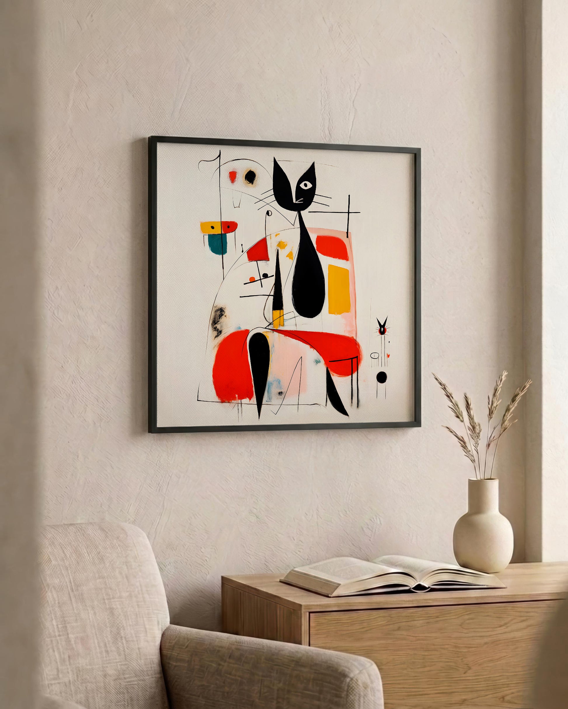 A framed abstract art print featuring a stylized black cat and geometric shapes in red, yellow, blue, and black on a white background, hanging on a textured beige wall above a wooden side table with an open book and a vase.